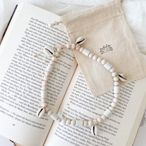 May include: A white and natural shell beaded necklace with white beads and gold accents. The necklace is on a book with the text "Aloha Dogs" on a small canvas bag.