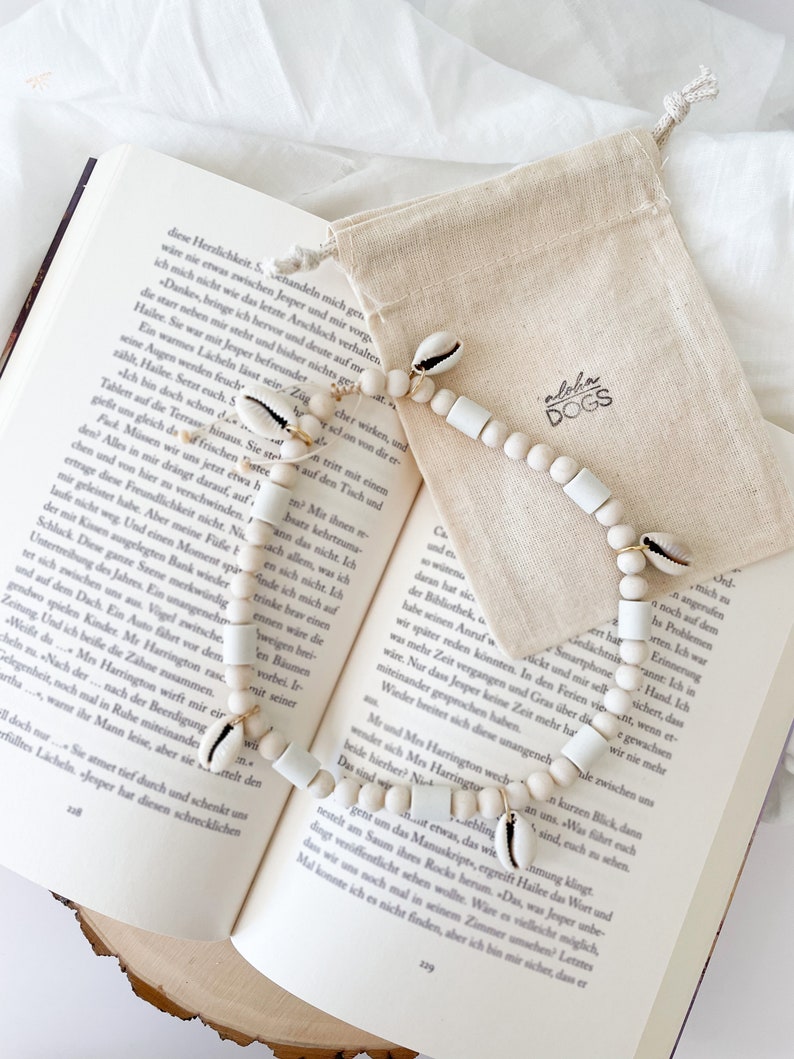 May include: A white and natural shell beaded necklace with white beads and gold accents. The necklace is on a book with the text "Aloha Dogs" on a small canvas bag.