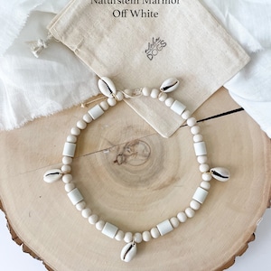May include: A white natural stone marble necklace with a shell charm. The necklace is on a wooden surface and comes with a white drawstring bag that says "Naturstein Marmor Off White" and "Aloha Dogs".