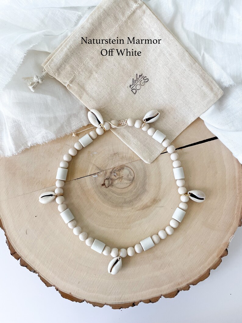 May include: A white natural stone marble necklace with a shell charm. The necklace is on a wooden surface and comes with a white drawstring bag that says "Naturstein Marmor Off White" and "Aloha Dogs".