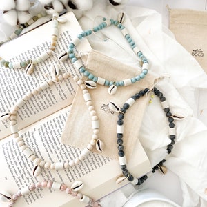 May include: Four necklaces made with white and natural colored beads, shells, and black lava rock. The necklaces are displayed on a book with the text "Aloha Dogs" on a small bag.