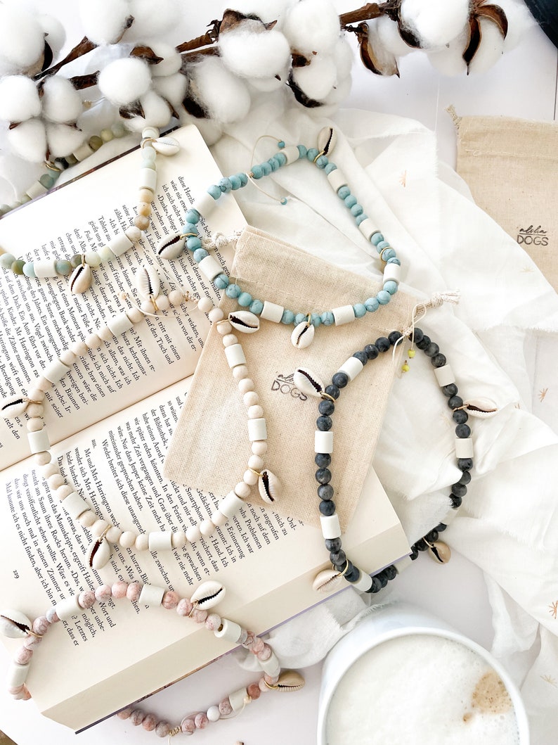 May include: Four necklaces made with white and natural colored beads, shells, and black lava rock. The necklaces are displayed on a book with the text "Aloha Dogs" on a small bag.