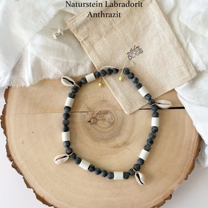 May include: A black and white beaded necklace with small seashells and a gold clasp. The necklace is on a wooden surface and a small white bag with the text "Aloha Dogs" is in the background. The text "Naturstein Labradorit Anthrazit" is also visible in the background.
