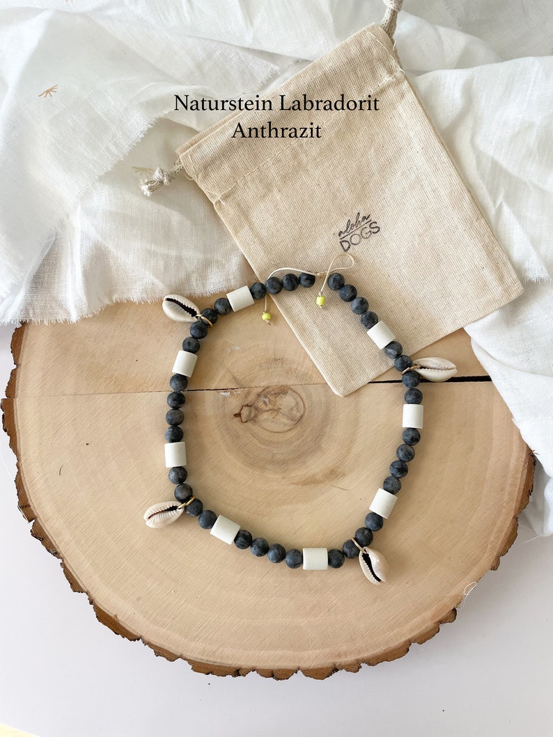 May include: A black and white beaded necklace with small seashells and a gold clasp. The necklace is on a wooden surface and a small white bag with the text "Aloha Dogs" is in the background. The text "Naturstein Labradorit Anthrazit" is also visible in the background.