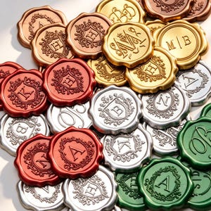 Puede incluir: Una colección de sellos de cera decorativos en varios colores, incluyendo dorado, plateado, bronce, rojo y verde. Cada sello presenta una letra o monograma diferente, rodeado de bordes ornamentados. Los sellos están dispuestos en un montón.