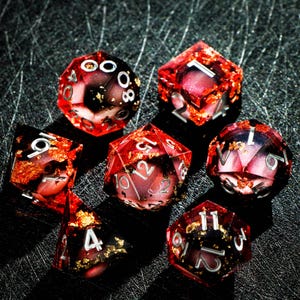 May include: Set of seven polyhedral dice for tabletop games. The dice are transparent red with gold flakes and white numbering. The dice include a d4, d6, d8, d10, d12, d20, and a percentile die.