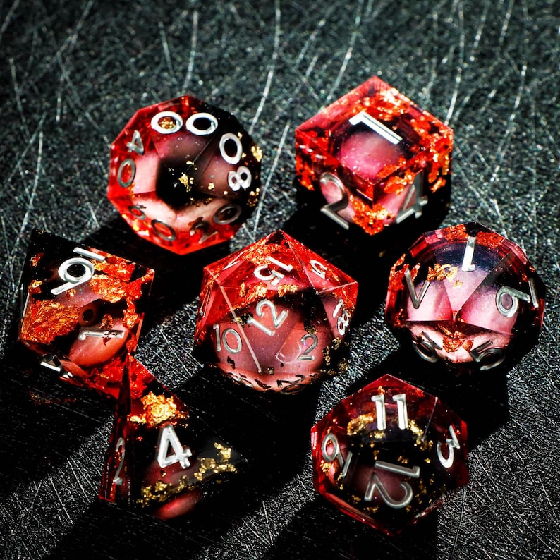 May include: Set of seven polyhedral dice for tabletop games. The dice are transparent red with gold flakes and white numbering. The dice include a d4, d6, d8, d10, d12, d20, and a percentile die.