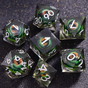 May include: A set of seven polyhedral dice made of clear resin, each containing a detailed green dragon eye with a vertical pupil and orange iris. The dice feature sharp edges and white engraved numbers, including a d20, d12, d10, d8, d6, and d4.