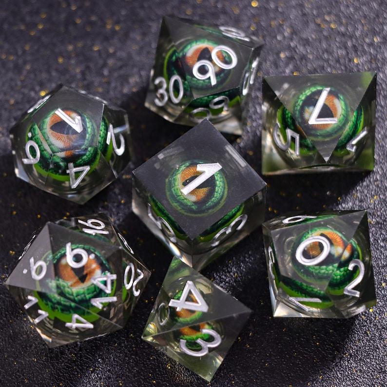 May include: A set of seven polyhedral dice made of clear resin, each containing a detailed green dragon eye with a vertical pupil and orange iris. The dice feature sharp edges and white engraved numbers, including a d20, d12, d10, d8, d6, and d4.