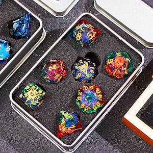 Puede incluir: Un conjunto de siete dados iridiscentes con motas doradas y números negros en una caja de metal plateada. Los dados son de varios colores, incluyendo rojo, azul, verde y morado.