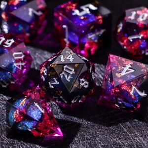 Peut inclure: Un ensemble de dés polyédriques à multiples facettes en résine translucide. Les dés présentent une base bleu et violet profond avec des éclats métalliques rouges et dorés incrustés. Chaque dé a des bords tranchants et des chiffres gothiques argentés.