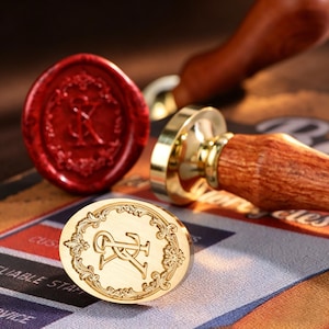 Może przedstawiać: Złoty metalowy stempel do laku z owalną powierzchnią, ozdobiony misternym filigranowym obrzeżem oraz wytłoczonymi, splecionymi literami R i K. Do stempla przymocowany jest drewniany uchwyt, a obok znajduje się czerwona pieczęć z tego samego wzoru.