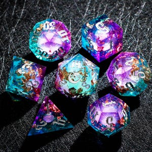 May include: Set of seven polyhedral dice in various shapes. The dice are made of clear resin with a mix of teal, purple, and gold leaf inclusions. Each die has silver numbering.