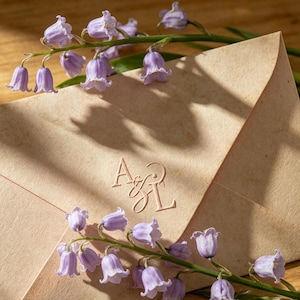 Puede incluir: Primer plano de un sobre beige con iniciales en relieve, adornado con delicadas flores moradas en forma de campana. Las flores están dispuestas alrededor del sobre, creando una estética suave y natural.