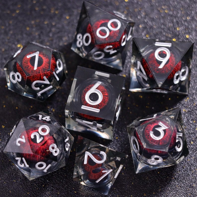May include: A set of seven polyhedral dice made of clear resin, each containing a realistic red iris and black pupil. The dice feature white engraved numbers, including a d20, d12, d10, d8, d6, d4, and a percentile die, all arranged on a dark surface.