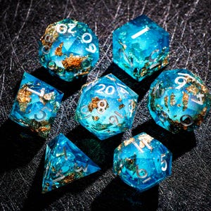 May include: A set of seven polyhedral dice for tabletop games. The dice are a translucent blue with gold foil inclusions and silver numbering. The dice include a d4, d6, d8, d10, d12, d20, and a percentile die.