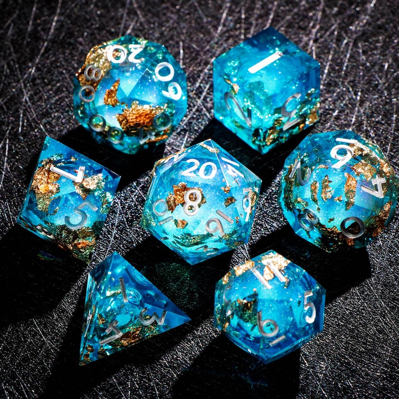 May include: A set of seven polyhedral dice for tabletop games. The dice are a translucent blue with gold foil inclusions and silver numbering. The dice include a d4, d6, d8, d10, d12, d20, and a percentile die.