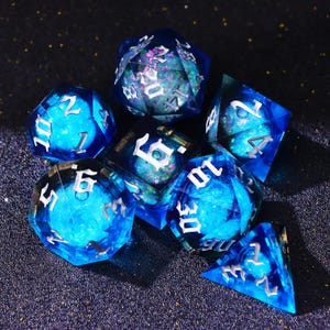 May include: A set of seven polyhedral dice in shades of blue with white numbering. The dice have a translucent, galaxy-like interior with glitter. The dice are designed for tabletop role-playing games.