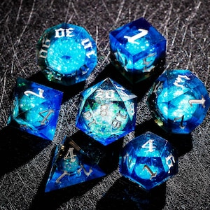May include: A set of seven polyhedral dice with a celestial theme. The dice are a translucent blue with a swirling, galaxy-like interior. Each die has silver numbering, perfect for tabletop gaming.
