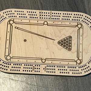 May include: Wooden cribbage board with a pool table design. The board features a central pool table graphic with a cue stick and a rack of billiard balls. The outer edges have holes for scoring. The board is made of light-colored wood.