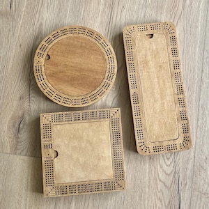 Peut inclure: Trois plateaux de cribbage en bois de formes circulaire, rectangulaire et carrée. Chaque plateau comporte un compartiment de rangement central avec un couvercle coulissant et des rangées de trous pour marquer les points. Le carré porte le texte START et FINISH.