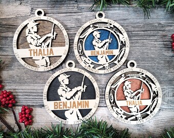 May include: Three round wooden ornaments with a fishing theme. Each ornament features a silhouette of a person fishing with a rod and reel. The ornaments are decorated with various fishing-related words and images, including "bait", "lake", "hook", "jig", "crank", "spinner", "spoon", "swivel", "reel", and "cast". The ornaments are personalized with the names "Thalia" and "Benjamin".