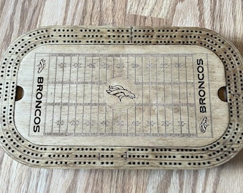 May include: Wooden cribbage board featuring a football field design with the Denver Broncos logo and team name. The board has multiple rows of holes for pegging and is made of light-colored wood.