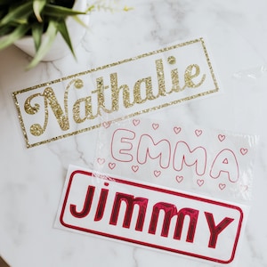 Op de afbeelding: Drie gepersonaliseerde naamstickers met gouden glitter, roze hartjes en een witte achtergrond. De namen zijn Nathalie, Emma en Jimmy.