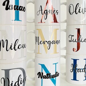 Op de afbeelding: Een verzameling witte keramische mokken met gepersonaliseerde namen en initialen in verschillende kleuren, waaronder goud, zilver, blauw en rood. De mokken zijn op elkaar gestapeld.