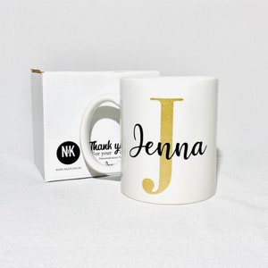 Pode incluir: Caneca de cerâmica branca com uma letra J de glitter dourado e o nome Jenna em escrita preta. A caneca está em uma caixa branca com o texto "Obrigado pela sua personalização" e um logotipo.