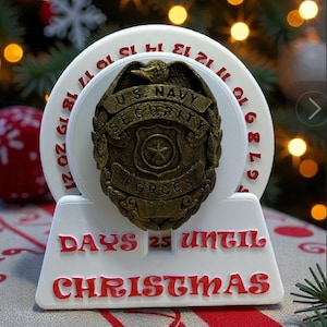 May include: A white and red Christmas countdown decoration. It features a US Navy Security Forces badge, a countdown display reading "25 Days Until Christmas", and a circular dial with numbers. The background includes a Christmas tree with bokeh lights.