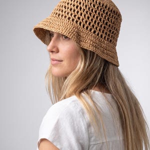 Puede incluir: Un sombrero de cubo marrón claro, de tejido abierto, con un ala ligeramente inclinada hacia abajo. El sombrero está hecho de un material tejido, probablemente paja o una fibra natural similar. El sombrero es de color neutro y tiene un estilo clásico e informal.