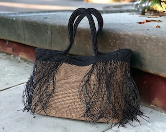 May include: A structured tote bag featuring a tan crocheted body and a contrasting black crocheted top border. The bag includes two black crocheted handles and long black fringe detailing that hangs from the upper section across the front.