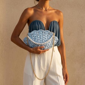 May include: A light blue crochet clutch bag shaped like a seashell, featuring a decorative strand of white faux pearls across the front and a gold-tone metal chain shoulder strap. The bag has a textured, fan-like pattern throughout its exterior.