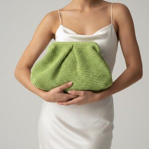 May include: A lime green, knitted clutch bag with a gathered top. The bag is held in front of a person wearing a white satin slip dress. The clutch has a soft, textured appearance.