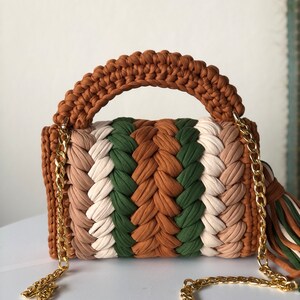 May include: A brown, green, white, and beige crocheted handbag with a gold chain strap and tassel.