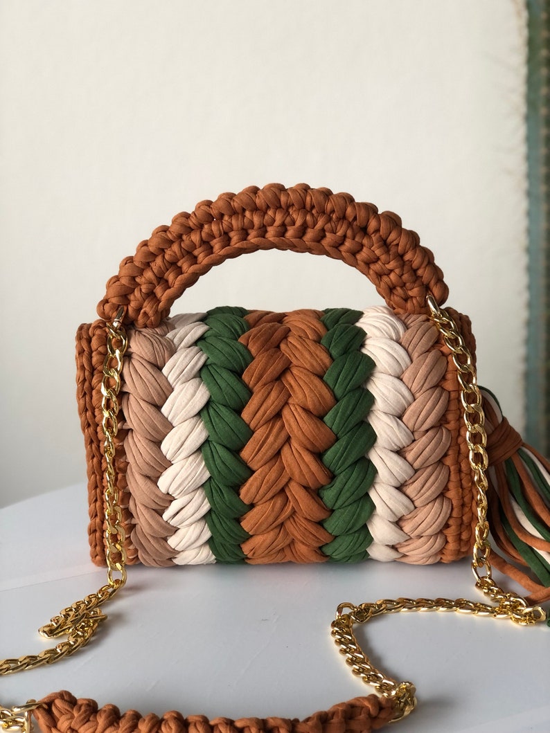 May include: A brown, green, white, and beige crocheted handbag with a gold chain strap and tassel.