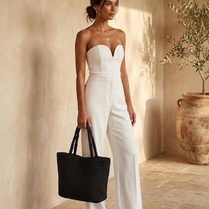 May include: A white strapless jumpsuit featuring a sweetheart neckline and wide-leg trousers, paired with a large black woven tote bag with two top handles. The jumpsuit has a fitted bodice and a clean, solid white finish throughout the garment.