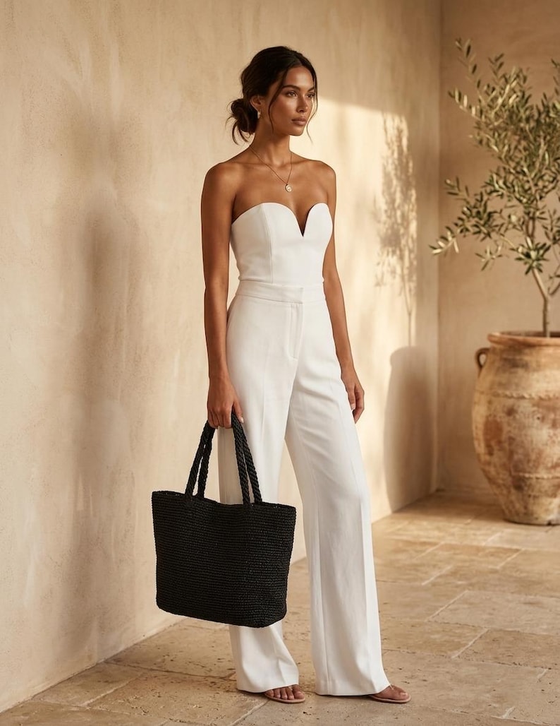 May include: A white strapless jumpsuit featuring a sweetheart neckline and wide-leg trousers, paired with a large black woven tote bag with two top handles. The jumpsuit has a fitted bodice and a clean, solid white finish throughout the garment.