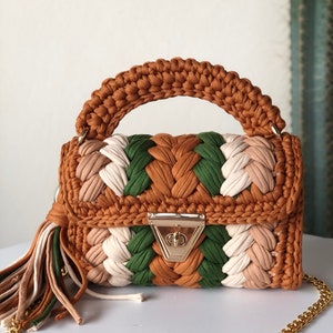 May include: A small, crocheted handbag with a brown, green, and white color scheme. The bag has a top handle and a gold chain strap. The bag has a gold clasp closure.