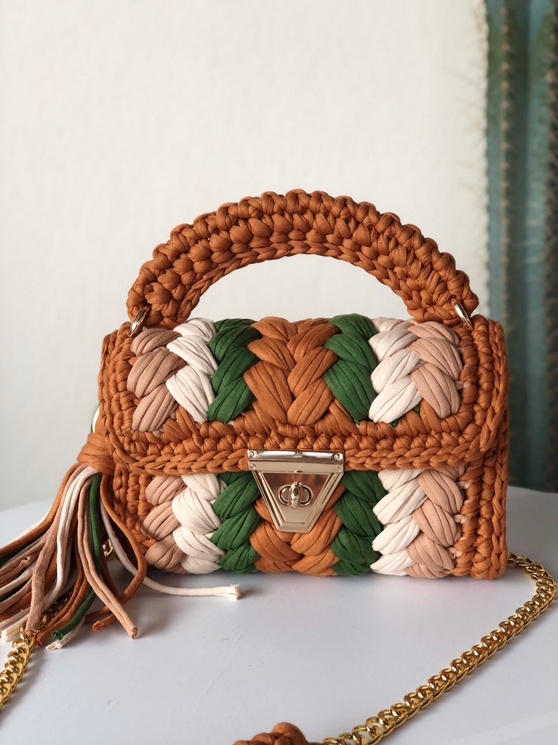May include: A small, crocheted handbag with a brown, green, and white color scheme. The bag has a top handle and a gold chain strap. The bag has a gold clasp closure.
