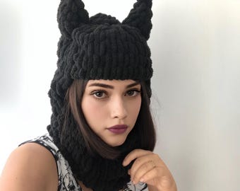 May include: A black chunky knit balaclava featuring two prominent pointed ear-like structures on top. The headgear is crafted from thick, soft yarn with a textured, looped stitch pattern that covers the head and neck area completely.