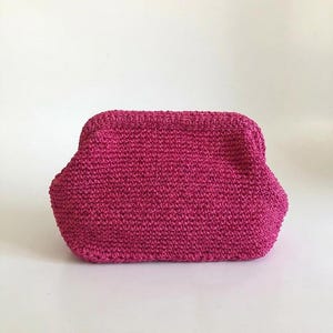 May include: A vibrant magenta crochet pouch featuring a structured frame closure. The accessory is crafted from a textured yarn with a consistent stitch pattern, showcasing a rounded rectangular shape with a wide base and a secure top opening.