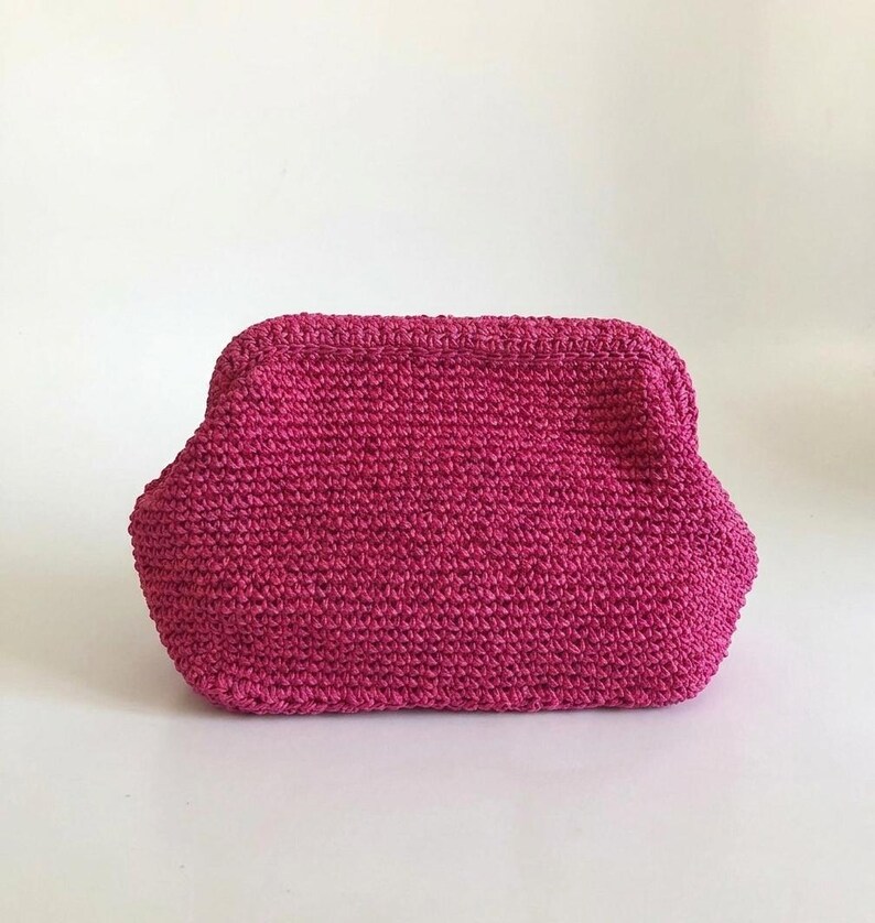May include: A vibrant magenta crochet pouch featuring a structured frame closure. The accessory is crafted from a textured yarn with a consistent stitch pattern, showcasing a rounded rectangular shape with a wide base and a secure top opening.