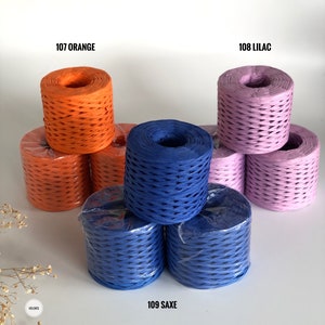 May include: Three spools of paper cord in orange, lilac, and blue. The spools are stacked on a white surface. The text "107 ORANGE", "108 LILAC", and "109 SAXE" is printed below each spool.