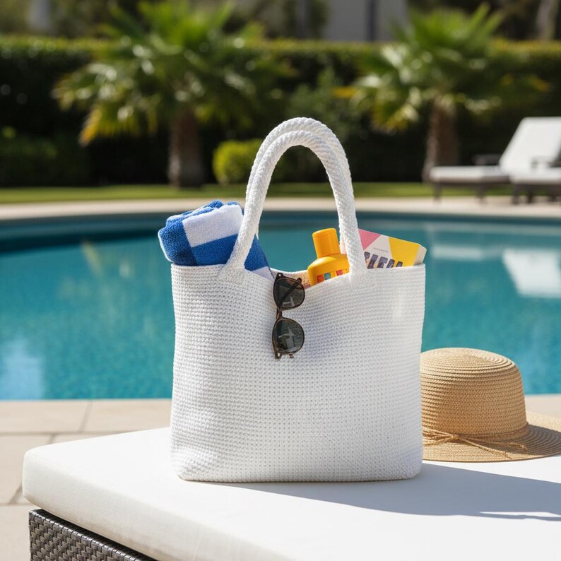 May include: A white woven tote bag with two sturdy handles. The bag contains a blue and white striped towel, a yellow sunscreen bottle, and a book. A pair of dark-rimmed sunglasses is hooked onto the front of the bag. A straw hat sits nearby.