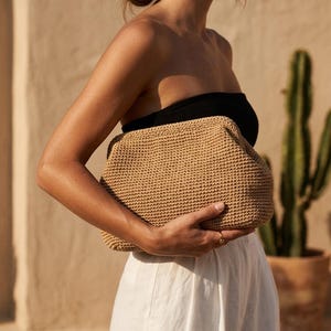Puede incluir: Un bolso de mano tipo sobre en color beige, tejido con una estructura r&iacute;gida y una superficie texturizada de nudos. El bolso presenta una forma rectangular con esquinas redondeadas y un cierre superior seguro, hecho de material natural.