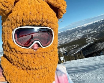May include: A mustard yellow chunky knit balaclava featuring two rounded ears on top, worn with white-framed ski goggles with dark reflective lenses. The balaclava covers the head and neck, leaving an opening for the eyes and nose.