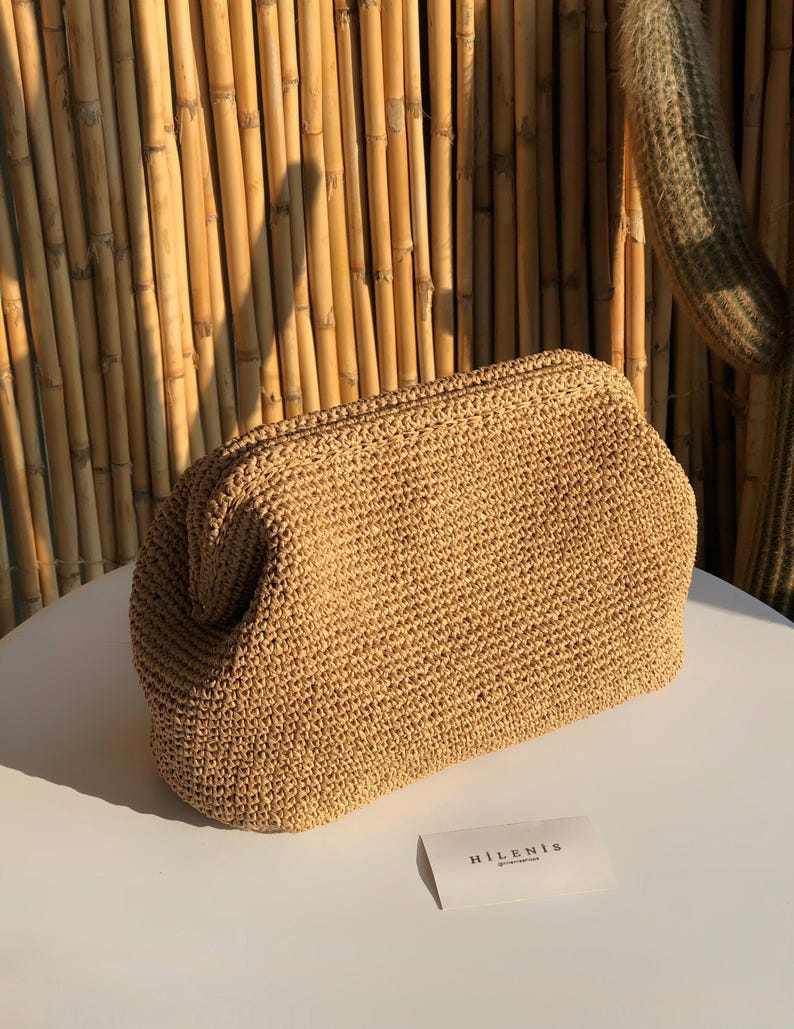 Puede incluir: Un bolso de mano de ganchillo en color beige con una abertura de marco estructurado. El bolso presenta un patr&oacute;n tejido y texturizado en todo su exterior. Una peque&ntilde;a tarjeta rectangular blanca con el texto HILENIS se sit&uacute;a frente al bolso.