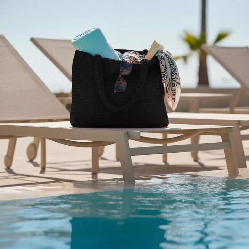 May include: A black woven tote bag with thick rope handles, containing a light blue towel, a pair of dark-rimmed sunglasses resting on the front, and a patterned white and black scarf draped over the side, all set against a bright outdoor background.